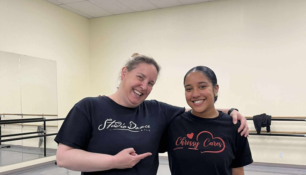 Two smiling women pose in a dance studio, one pointing to the other's black shirt reading "Chrissy Cares".