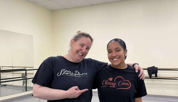 Two smiling women pose in a dance studio, one pointing to the other's black shirt reading "Chrissy Cares".