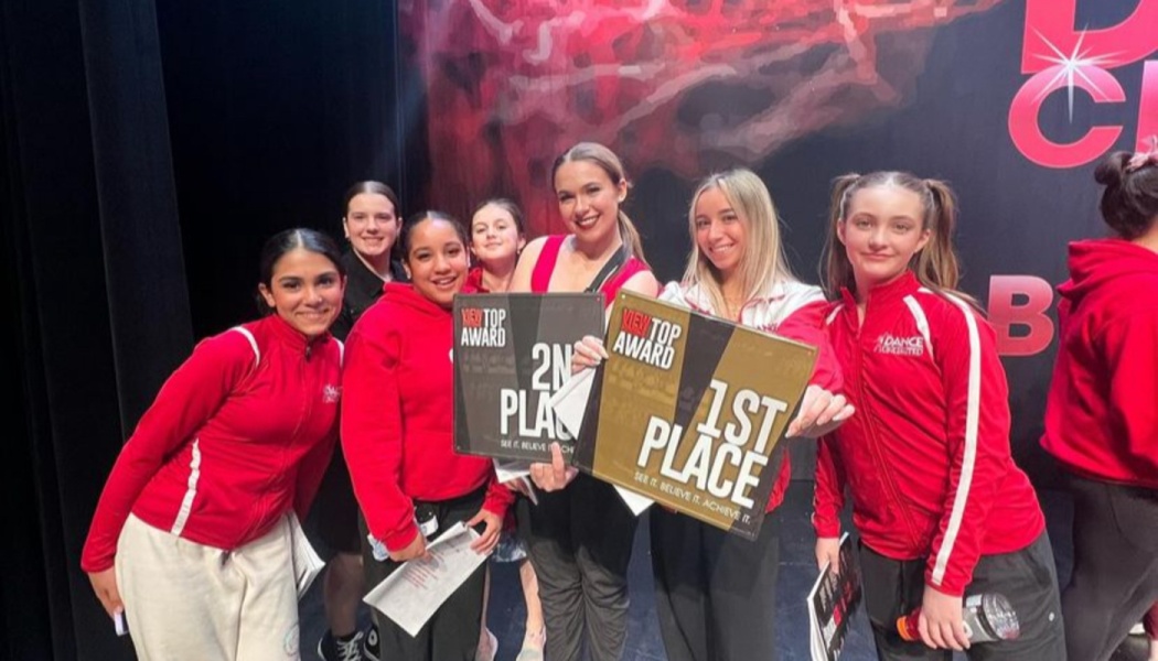 Smiling young dancers in red tracksuits proudly holding 1st and 2nd place competition award plaques.