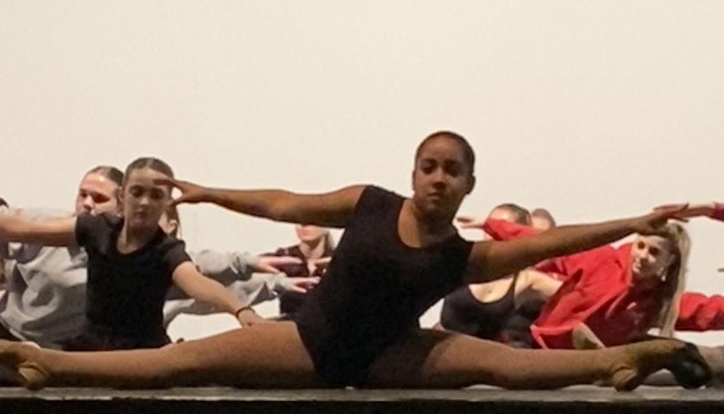 A dancer in a black leotard performs a floor split on stage alongside her ensemble.