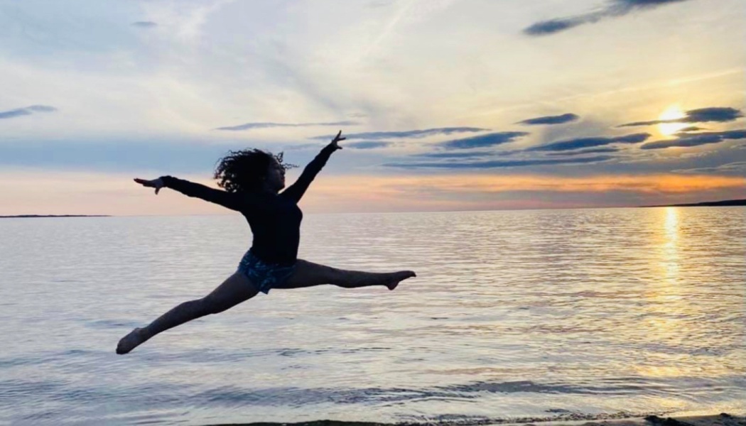 A dancer silhouetted mid-air performing a split leap on the beach during a sunset.