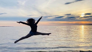 A dancer silhouetted mid-air performing a split leap on the beach during a sunset.