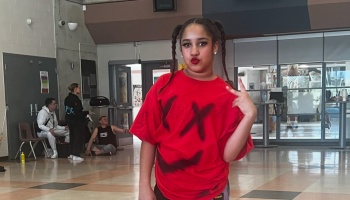 Young female dancer in pigtails making a peace sign, wearing a red shirt with a black spray-painted face.