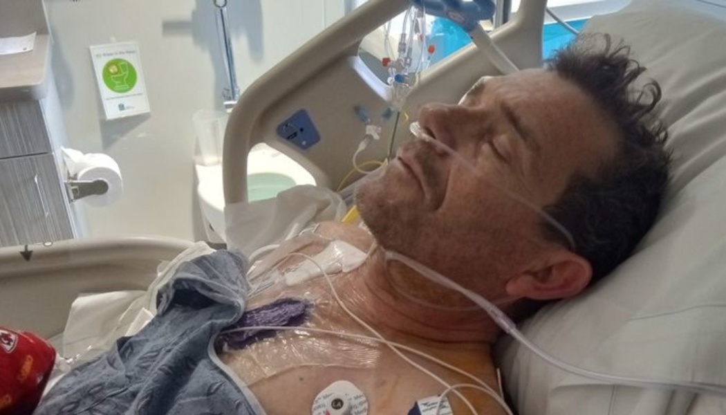 A middle-aged man lies in a hospital bed with a nasal cannula and medical sensors on his chest.
