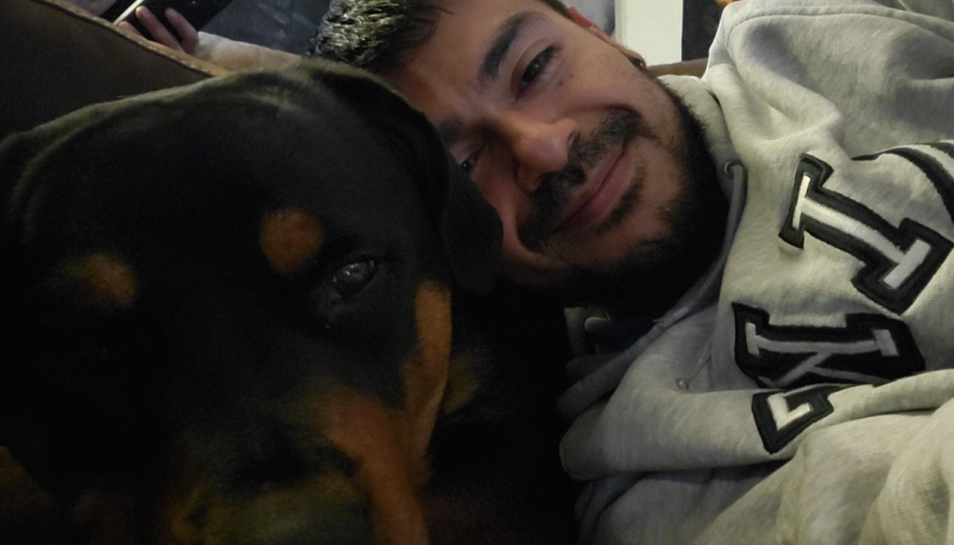 A smiling man in a grey hoodie cuddles closely next to a black and tan Rottweiler.
