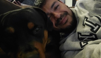 A smiling man in a grey hoodie cuddles closely next to a black and tan Rottweiler.