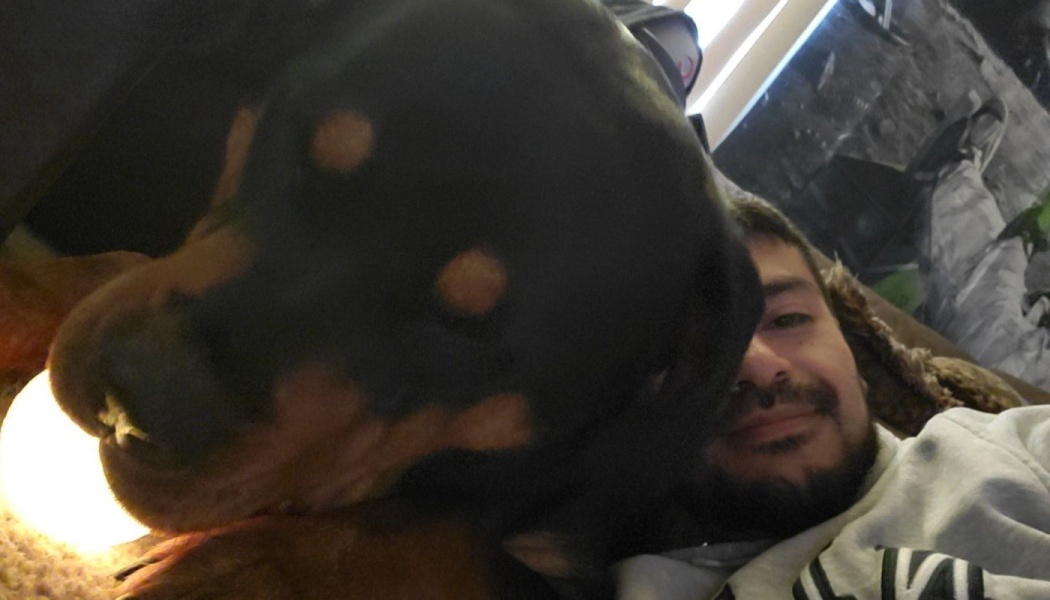 A smiling bearded man lying down beside a large black and tan dog leaning close to the camera.