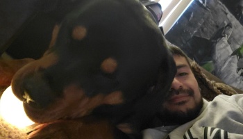 A smiling bearded man lying down beside a large black and tan dog leaning close to the camera.