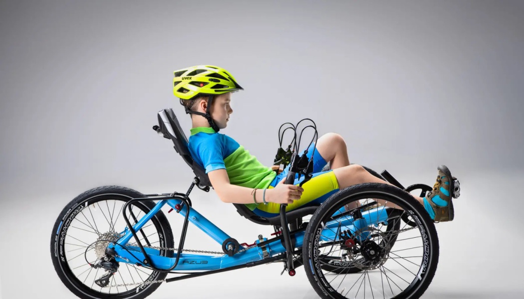 A young boy wearing a yellow helmet seated on a bright blue adaptive recumbent tricycle.