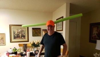 A smiling man in an orange cap playfully balances a long green pool noodle on his head.