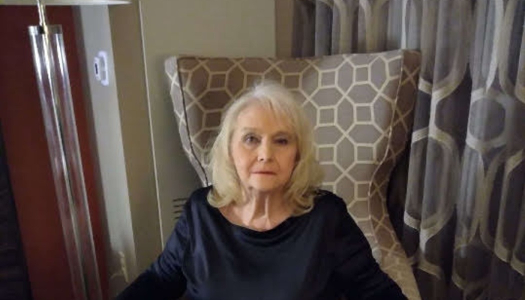 An elderly blonde woman in a black shirt sitting in a patterned high-back armchair.