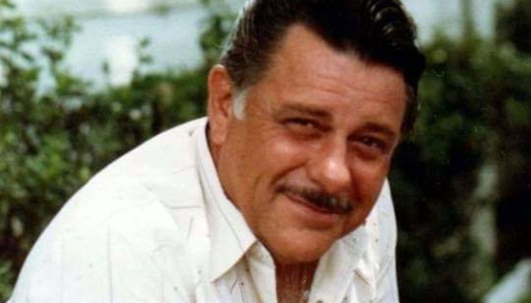 Middle-aged man with a mustache and dark hair smiling softly in a white collared shirt with greenery behind him.
