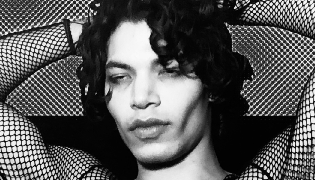 Black and white portrait of a person with dark curly hair in fishnet sleeves posing with hands behind their head.