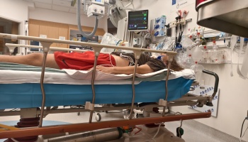 A patient in a neck brace lies on a hospital stretcher surrounded by medical equipment and monitors.