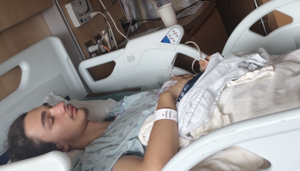 A young man rests in a hospital bed with an IV line attached to his hand and an ID bracelet on his wrist.