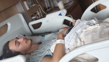 A young man rests in a hospital bed with an IV line attached to his hand and an ID bracelet on his wrist.