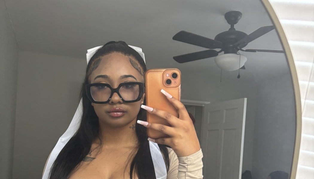 A young woman with black glasses and long manicured nails taking a mirror selfie with her smartphone.