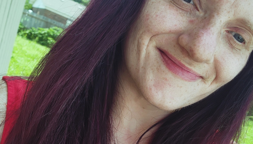 Close-up selfie of a woman with long dark purple hair, freckles, and a gentle smile outdoors.