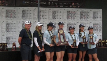 A women's golf team and coaches wearing leis pose with NCAA trophies in front of a scoreboard.