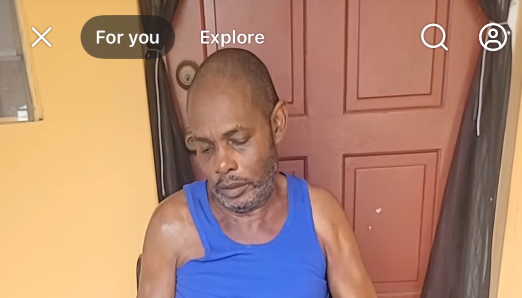 A man in a blue tank top looking down with a somber expression in front of a red door.