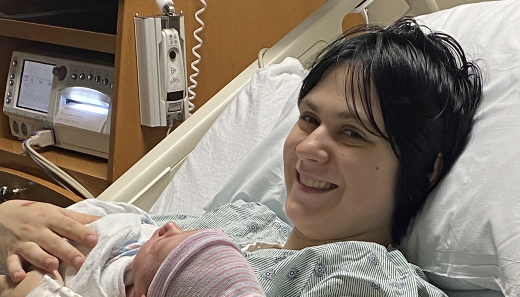A smiling mother in a hospital gown lies in bed holding her swaddled newborn baby.