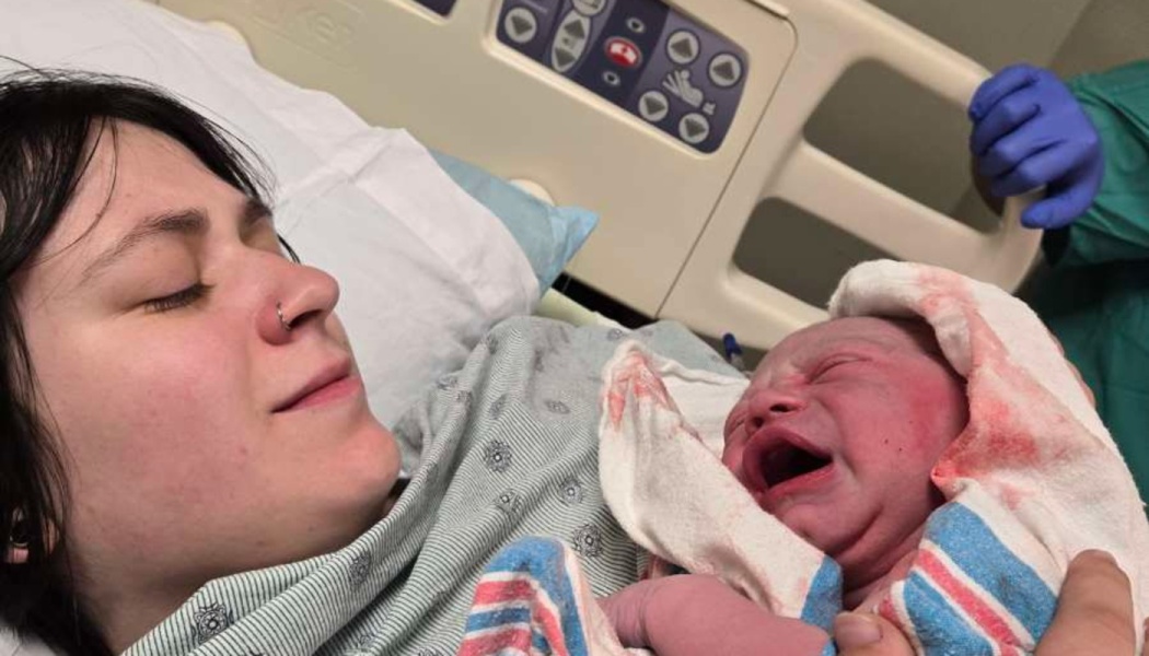 A mother lying in a hospital bed looks lovingly at her newborn baby wrapped in a hospital blanket.