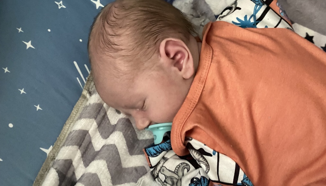 A newborn baby sleeps peacefully with a light blue pacifier, resting on a patterned blanket.