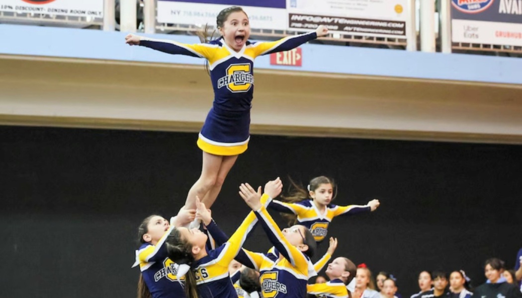 A young cheerleader shouts with excitement as her team lifts her into the air during a stunt routine.