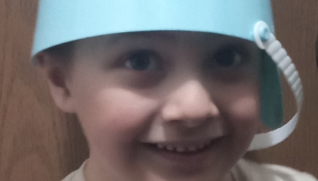 A smiling young child wearing a light blue plastic bucket like a helmet.