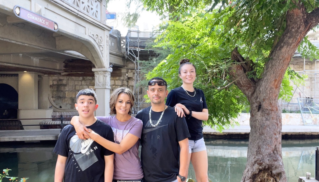 A family of four smiling together outdoors near a riverwalk with a stone bridge in the background.