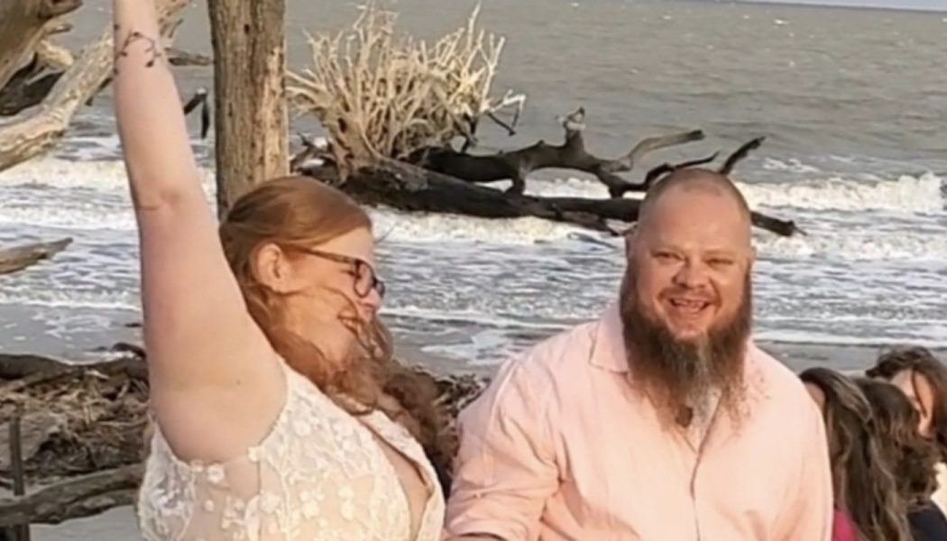 A happy woman in a white lace dress raises her arm next to a smiling bearded man on a beach with driftwood.