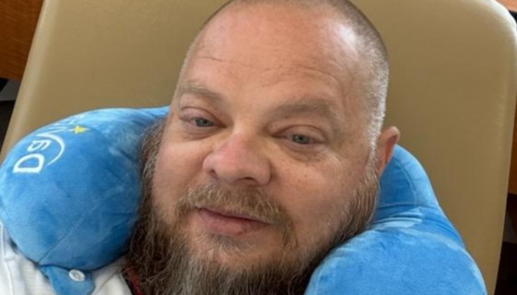 A bearded man with a shaved head rests in a chair wearing a bright blue neck pillow.