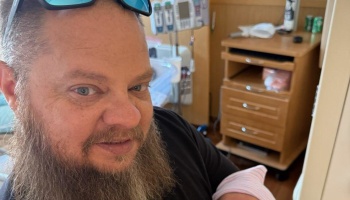 A smiling bearded man holding a swaddled newborn baby in a hospital room.