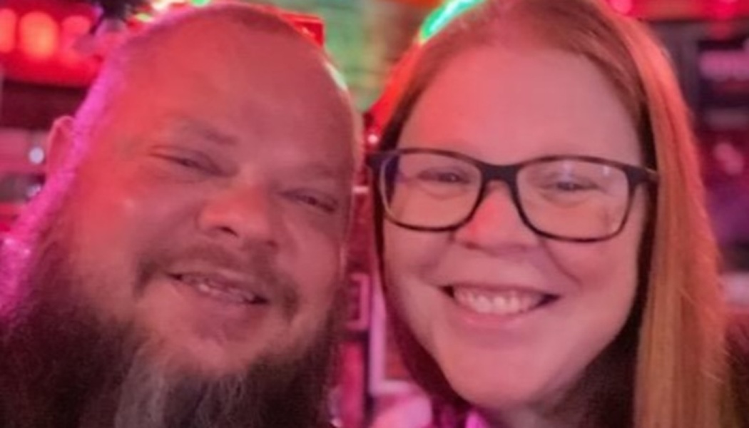 A smiling bearded man and a woman with red hair and glasses pose together under ambient red lighting.