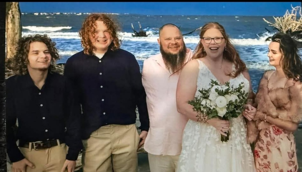 A bride holding a bouquet poses with her groom and family on a beach with ocean waves behind them.