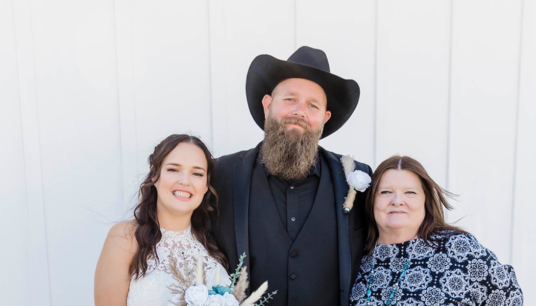 A bride in a white dress, a bearded groom in a black cowboy hat, and an older woman pose together smiling.