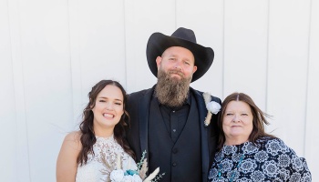 A bride in a white dress, a bearded groom in a black cowboy hat, and an older woman pose together smiling.