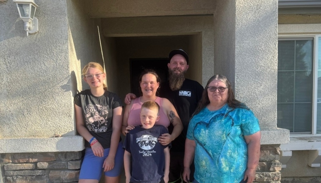 A smiling family of five standing together outside on the front porch of their home.