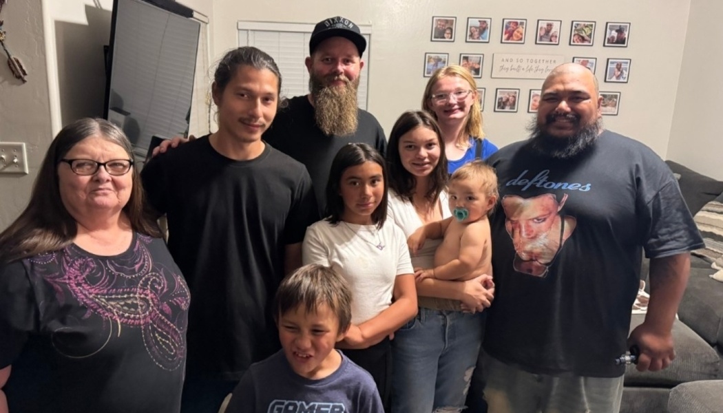A smiling multi-generational family of nine adults, teens, children, and a baby posing together indoors.