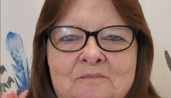 Close-up portrait of a woman with reddish-brown hair and black glasses looking at the camera.