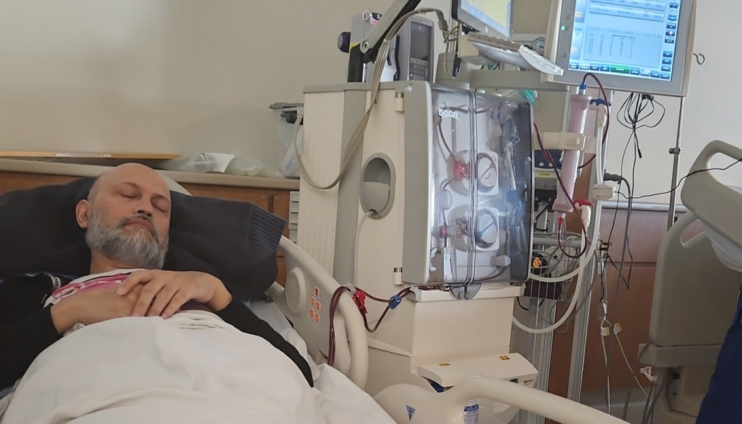A man with a gray beard rests in a hospital bed beside a dialysis machine.