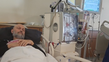 A man with a gray beard rests in a hospital bed beside a dialysis machine.
