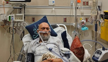 A bearded man rests in a hospital bed with an oxygen tube, surrounded by medical equipment.
