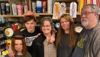 A smiling family of five poses together inside a store, with two younger members wearing fun hats.