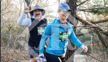 Two cheerful trail runners wearing race bibs smile as they jog along a wooded path.