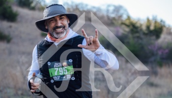 Smiling bearded trail runner wearing a sun hat and race bib, making a hand gesture and holding a water bottle.