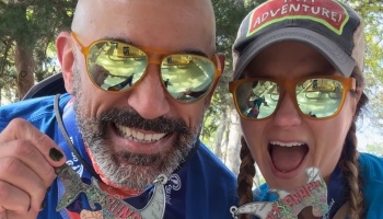 A smiling man and excited woman wearing sunglasses hold up their shark-shaped finisher medals.