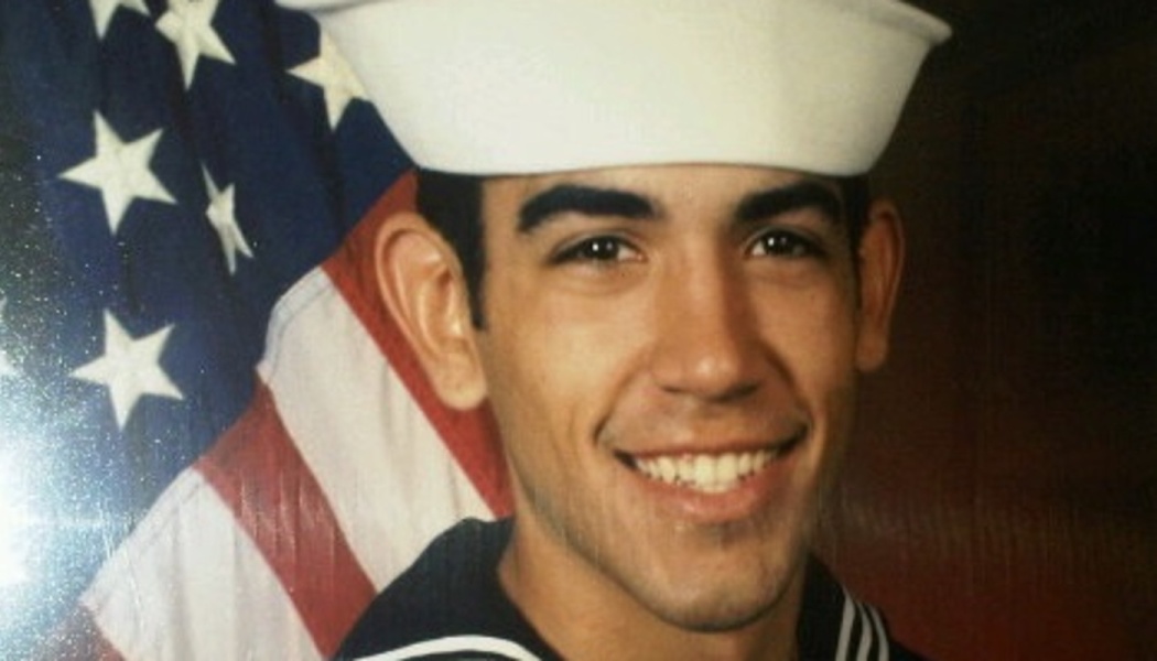 A smiling young US Navy sailor in uniform posing in front of an American flag.