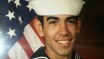 A smiling young US Navy sailor in uniform posing in front of an American flag.