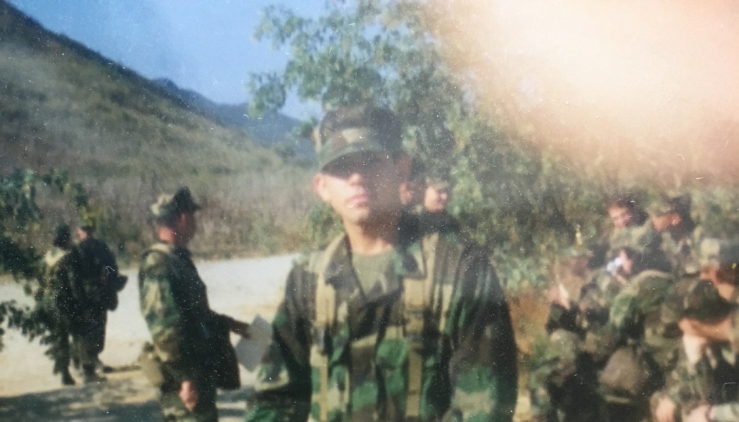 A young soldier in military camouflage uniform stands outdoors with other soldiers in a hilly landscape.